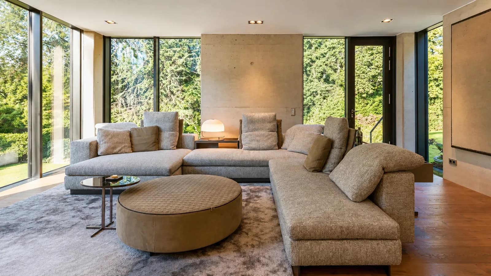 Wohnzimmer mit großer Polsterlandschaft vor Sichtbetonwand und raumhoher Verglasung zum Garten, Hartenfels