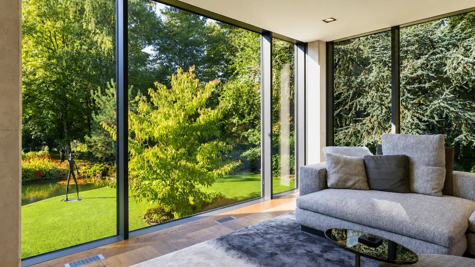 Verglaste Wohnecke mit Sofa und Blick auf den Gartenteich mit Skulptur, Hartenfels